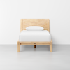 A single wooden bed with a light wood frame, white bedding, and one pillow, placed against a plain white wall on a smooth gray floor.