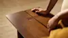 A person rests their hands on a smooth, brown wooden table with a narrow rectangular slot in the center. The background is softly blurred.