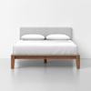 A modern bed with a light gray upholstered headboard, white bedding, and a wooden frame, set against a plain white wall and floor.