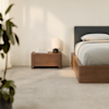A modern bedroom with a wooden bed frame and nightstand, white bedding, a lamp, and a small plant on the carpeted floor. Soft natural light enters from a window with sheer curtains.