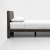 A minimalistic wooden bed frame with a padded headboard, neatly made with white bedding and a pillow, sits against a plain white wall on a smooth white floor.
