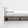 A minimalist side view of a modern bed with a light fabric headboard, white bedding, and a simple wooden frame, set against a plain white wall and floor.