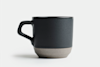 A black ceramic coffee mug with a matte finish and a contrasting light gray textured base, set against a plain white background.