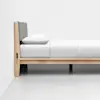 A minimalist wooden bed with a light-colored frame, a gray upholstered headboard, and neatly arranged white bedding is positioned against a plain white wall in a bright room.