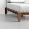 A close-up view of a wooden bed frame with clean lines and tapered legs, featuring a neatly made bed with white sheets and a pillow, set on a light gray floor.