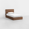 A single wooden bed with a simple headboard, dressed in white sheets and a white pillow, placed in a minimal, empty room with white walls and a light gray floor.