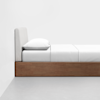 A minimal, modern bed with a light gray upholstered headboard, white bedding, and a wooden base, placed against a plain white wall on a smooth gray floor.