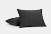 Two black square pillows with a subtle grid pattern on the surface, placed on a plain white background. One pillow is upright while the other lies flat behind it.