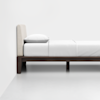 A minimalist bed with a beige upholstered headboard, a white pillow, and white bedding on a dark wooden frame, placed against a plain white wall in a modern, uncluttered room.