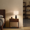 A bedside table with a lit lamp, a small cup, glasses, and a book on the lower shelf, positioned next to a wooden bed frame.