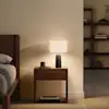 A bedside table with a lit lamp, a small cup, glasses, and a book on the lower shelf, positioned next to a wooden bed frame.