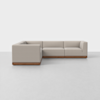 A modern L-shaped sectional sofa with light beige upholstery and a wooden base, positioned in a minimal, bright room with a plain white background and a smooth, light-colored floor.