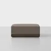 A square, brown upholstered ottoman with a dark wooden base, placed on a light gray floor against a plain white background.