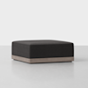 A modern, square ottoman with a dark upholstered cushion and a light wooden base, placed on a light, smooth floor against a plain white background.