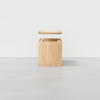 A light wooden box with a square shape is shown with its lid floating above, revealing an empty interior. The box sits on a smooth gray surface against a plain white background.