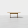A round, light wood coffee table with three legs, placed on a plain, light gray floor against a white background.