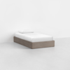 A minimalist bed with a light wooden frame, white mattress, white sheet, and a single white pillow, placed in an empty, bright room with a light gray floor and plain white walls.