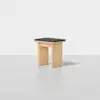 A minimalist wooden stool with a rectangular black cushioned seat, set against a plain white background. The stool has wide, flat legs and a simple, modern design.