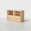 A minimalist wooden bookshelf with two open shelves on top and four closed drawers below, placed on a light gray floor against a plain white background.