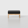A minimalist wooden stool with a rectangular black cushioned seat, set against a plain light gray background.