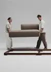 A woman and a man lift a large, brown sofa cushion together, positioning it onto a wooden sofa frame on a plain, light background.
