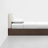 A minimalist bed with a white mattress, white pillow, and white bedding, set on a dark wooden frame against a plain white wall and a light gray floor.