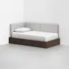 A modern daybed with a dark wooden base and light gray upholstered back and side panels, placed in the corner of a white, minimalistic room. The bed is neatly made with white bedding and a single pillow.