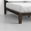 Close-up view of a modern wooden bed frame with white bedding, showing the mattress and sturdy, dark wood legs on a smooth, light-colored floor.
