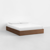A minimalist bed with a wooden platform frame, white mattress, and two white pillows, set against a plain white wall and floor.