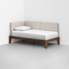 A minimalist daybed with a wooden frame, white mattress, and beige upholstered back and side panels, positioned in the corner of a plain, light-colored room.