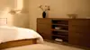 A modern, low wooden sideboard with drawers stands against a beige wall. On top are a round lamp, potted plant, books, ceramic vases, and decorative objects. Part of a neatly made bed is visible in the foreground.