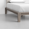 A close-up view of the corner of a modern bed with light wood frame and white bedding, positioned on a smooth, light-colored floor against a plain white wall.