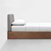 A minimalist bed with a wooden frame, gray upholstered headboard, white pillow, and white bedding, placed against a plain white wall on a light gray floor.