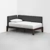 A modern daybed with a dark wooden frame, white mattress, single white pillow, and dark gray upholstered back and side panels, placed in a corner of a minimalist room with white walls and a light gray floor.