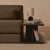 A modern wooden side table beside a brown sofa holds a stack of magazines, a small black lamp with a white shade, and several books on the lower shelf, all placed on a light-colored rug and hardwood floor.