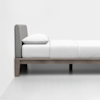 A neatly made single bed with a gray upholstered headboard, white pillow, and white bedding on a wooden frame, positioned against a plain white wall in a minimalistic room.