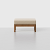 A modern, minimalist ottoman with a beige cushioned top and a wooden base, set against a plain light gray background.