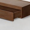 A close-up of a wooden storage bed with a partially open drawer, showing smooth brown wood and clean, modern lines on a light gray floor.