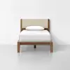 A single wooden bed with a light beige upholstered headboard, white bedding, and a single white pillow, positioned against a plain white wall on a light-colored floor.