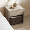 A modern bedside table with a light stone top, drawer, and open shelf holding a grey fabric basket. On top are a small vase with a single twig and a round dish with two wooden items.