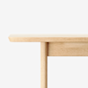 Close-up view of the corner of a light wood table against a white background, showing the tabletop edge and one supporting leg.