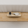 A wooden tray on a light wooden surface holds a black cup on a saucer, an open notebook, and a pen, with a white bed in the background.