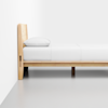 A minimalist wooden bed frame with a light finish, white bedsheet, and a single pillow, positioned against a plain white wall on a light gray floor.
