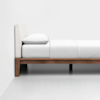 A minimalist bed with a wooden frame, white upholstered headboard, white pillow, and neatly made white bedding, positioned against a plain white wall and floor.