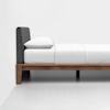 A modern bed with a dark gray upholstered headboard, white pillow, and white bedding on a wooden frame, placed against a plain white wall and floor.