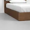 A close-up view of the corner of a wooden bed frame with a white mattress, white pillow, and white bedding, placed on a smooth gray floor.