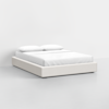 A modern, low-profile bed with a light beige upholstered frame, white mattress, and two white pillows, set against a plain white background.