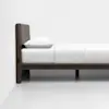 A minimalist wooden bed with a padded headboard, white bedding, and a single white pillow, set against a plain white wall and floor.