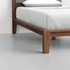 Close-up of a wooden bed frame with clean lines and tapered legs, featuring a white mattress and bedding. The floor is light gray and the background wall is white, creating a minimalist look.