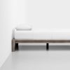 A minimalist bed with a wooden frame, white bedding, and a single pillow, placed against a plain white wall on a smooth, light gray floor.